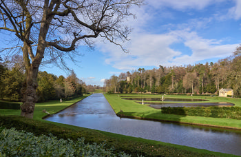 This landscape photograph captures a scenic view of the River Skell as it flows through Studley Royal Park in North Yorkshire, England, in the late afternoon during early spring. The image showcases the tranquil river bordered by well-manicured lawns and classical statues, with a clear, sunny sky illuminating the lush surroundings. In the middle distance, the formal water gardens and elegant structures typical of Studley Royal Park, a UNESCO World Heritage Site, are visible, set against a backdrop of tall trees, some still bare from winter. The scene highlights the historic beauty of this part of the United Kingdom, emphasizing the harmonious relationship between nature and designed landscape that characterizes this renowned location.